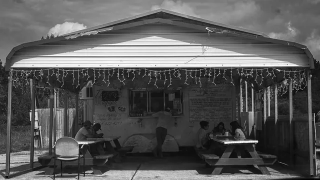 Black-and-white photo of Taqueria Raquel, a roadside taco stand in Jasper, Alabama, with diners at picnic tables under string lights